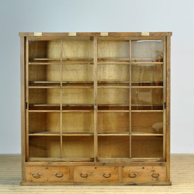 Japanese wooden cabinet featuring sliding glass doors, multiple shelves, and three drawers at the base. The cabinet bears...