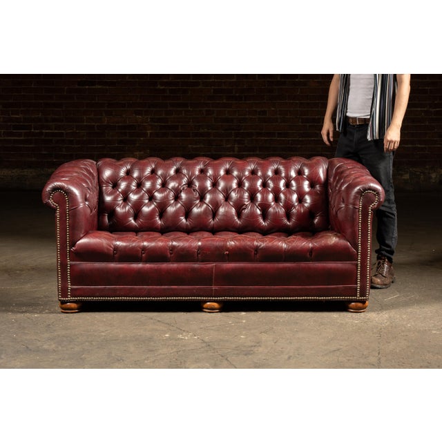 Classic tufted leather Chesterfield sofa in a deep burgundy tone with rolled arms, buttoned back, and brass nailhead trim....
