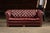Classic tufted leather Chesterfield sofa in a deep burgundy tone with rolled arms, buttoned back, and brass nailhead trim....