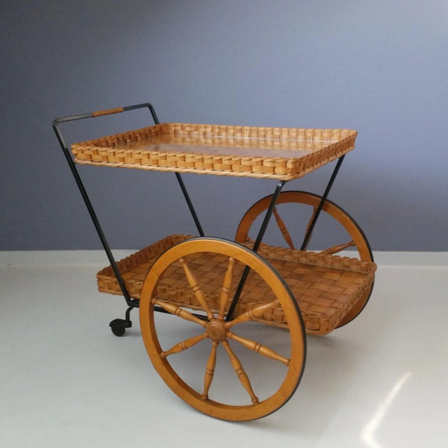 This serving trolley features a black metal frame and two tiers of woven beech bark. The big wheels are made of wood and...