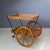 This serving trolley features a black metal frame and two tiers of woven beech bark. The big wheels are made of wood and...