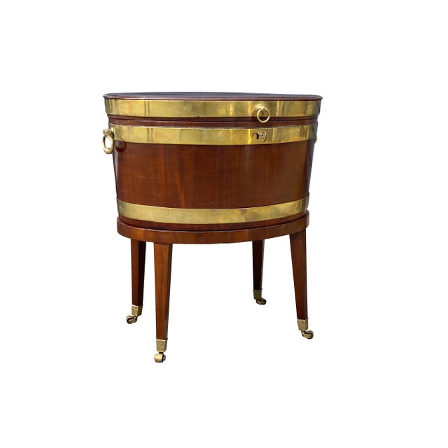 Oval hinged top over a conforming brass strapped case with brass handles, raised on square tapered legs with casters. Ex...