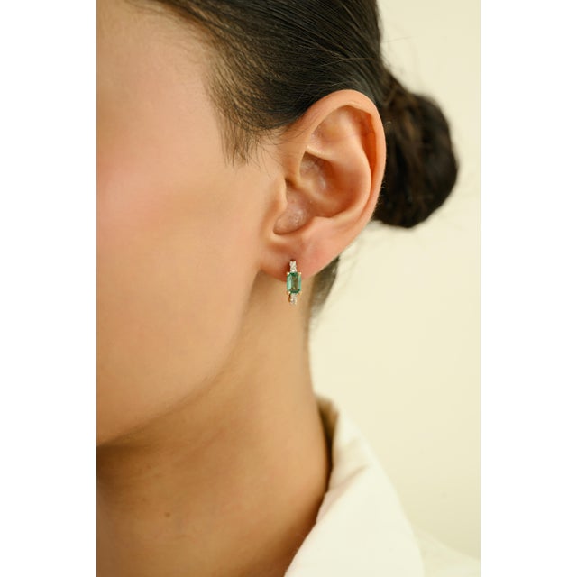 This elegant pair of huggie hoop earrings features 2 emerald cut emeralds totaling 1.02 carats, set in rich 14k yellow...