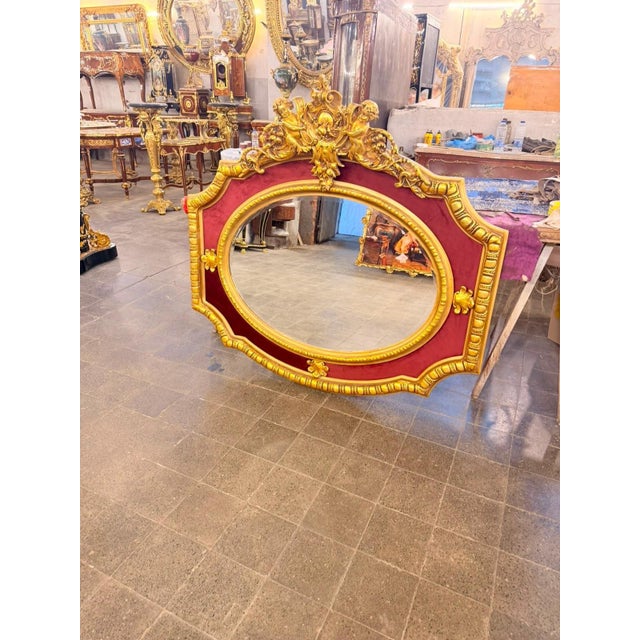French Rococo-style statement mirror featuring a richly sculpted gilt frame, deep burgundy inset panels, and an elaborate...