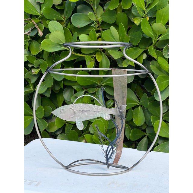 A sculptural metal “fish bowl” featuring a textured fish suspended within an open wire vessel, surrounded by hand-formed...