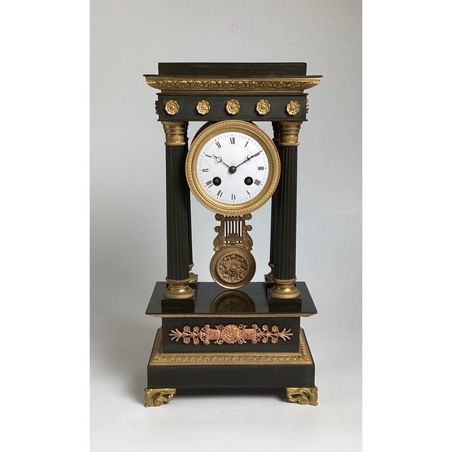An elegant neoclassical bronze portico clock. The patinated bronze with gilt bronze accents with round white porcelain...