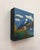 This encaustic work on wood panel captures a serene landscape, featuring a charming cottage nestled among rolling hills...