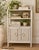Charming vintage white wicker étagère featuring a gracefully curved frame, woven shelving, and a double-door storage...