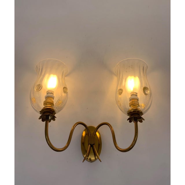 This pair of two-light wall lamps features a polished brass frame, featuring a central oval base from which two curved,...