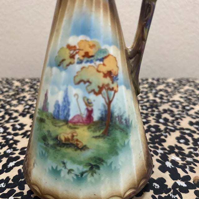 This elegant porcelain pitcher, or ewer, showcases a finely detailed, hand-painted pastoral landscape featuring a woman in...