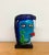 A whimsical sculptural art glass face composed in vivid shades of turquoise, cobalt blue, green, red, white, and brown....