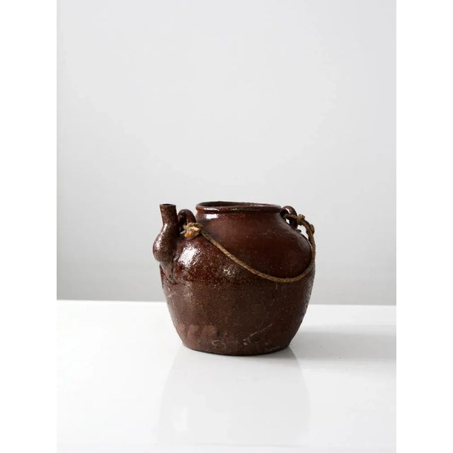 This antique clay bean pot with a brown Albany slip is from the late 19th century. It has a small spout and a braided rope...