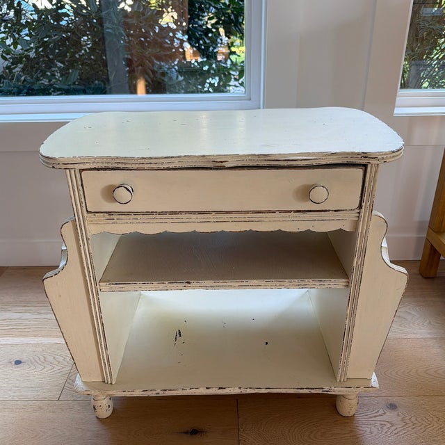 Charming vintage nightstand/side table featuring a creamy ivory painted finish, softly rounded top, and whimsical...