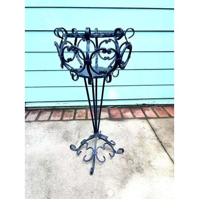 A tall vintage metal plant stand featuring an open scrollwork basket, slender tripod-style supports, and a decorative...