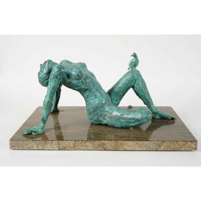 An alluring patinated bronze sculpture of a nude woman laying on a stone plinth; she is facing the sky and is accompanied...