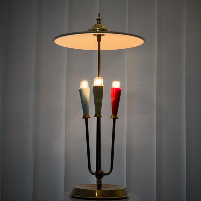 A modernist table lamp crafted from brass, featuring three arms set on a circular base. Each arm holds a conical...