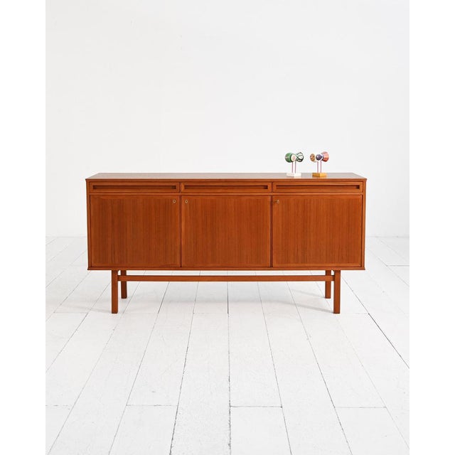 Teak sideboard with three hinged doors, three upper drawers, and a raised structure on linear legs. Scandinavian...