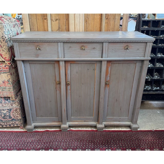 This three-door buffet features classic Gustavian-style reeded detailing across the cabinet doors and drawer fronts. The...