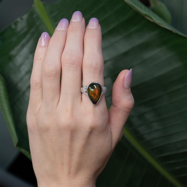 A striking 7.5ct Black Opal takes center stage, flashing vivid play-of-color against a deep, dramatic base. Framed by...