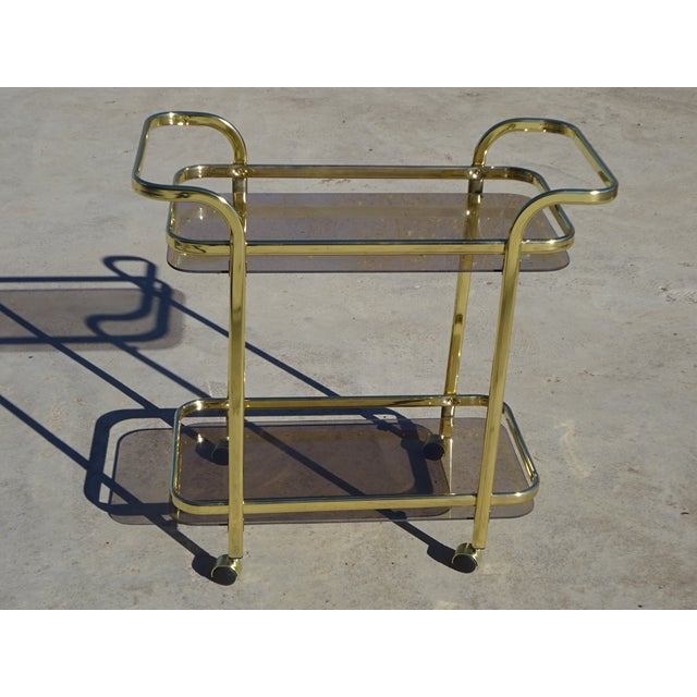 Gold-tone metal serving cart with a flattened tubular frame and smoked glass shelves. Two tempered glass shelves framed by...