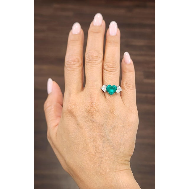 A captivating 3.11-carat GIA-certified Colombian heart-cut emerald ring, flanked by two GIA-certified natural dazzling...