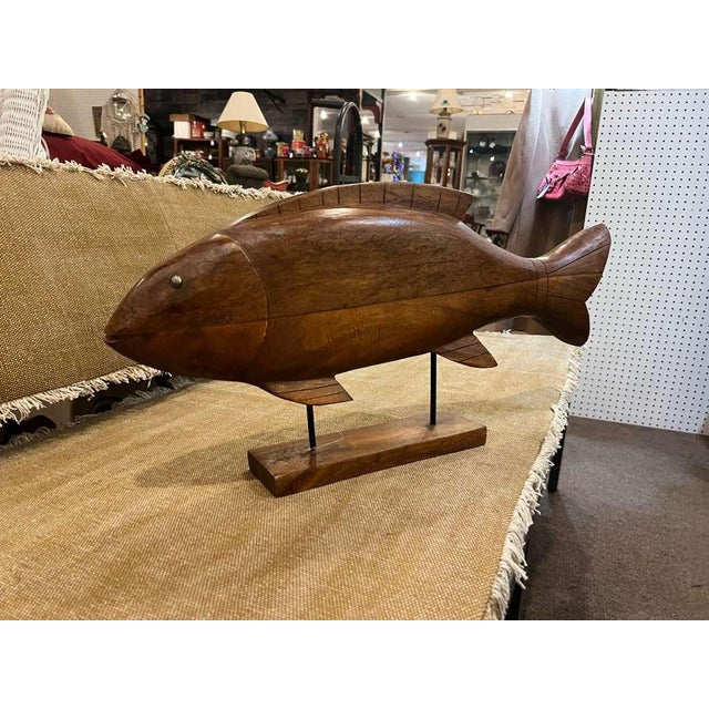 A beautifully crafted carved wooden fish statue mounted on a stand, made circa 2000s in the Coastal / Nautical style. The...