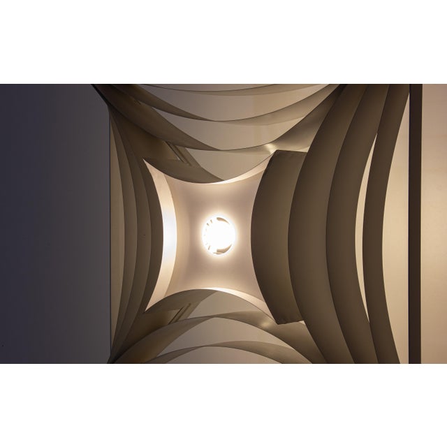 Square metal frame with curved internal elements forming a layered structure around a central light source. The lamp emits...