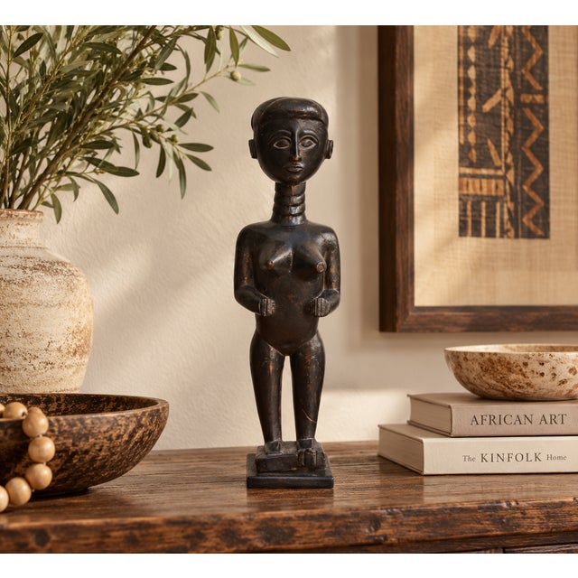 A slim standing woman carved in dark wood and polished to a deep, glossy black. She has an elongated, ringed neck, a close...