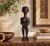 A slim standing woman carved in dark wood and polished to a deep, glossy black. She has an elongated, ringed neck, a close...