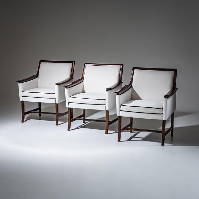 Set of three armchairs, model “Minerva,” with dark-stained wooden frames. The chairs feature slightly flared armrests and...