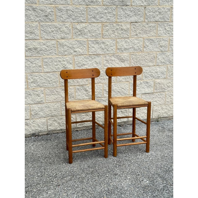 Pair of wood bar stools with woven rush seats. Each stool has a curved horizontal back rail, round legs, and lower...