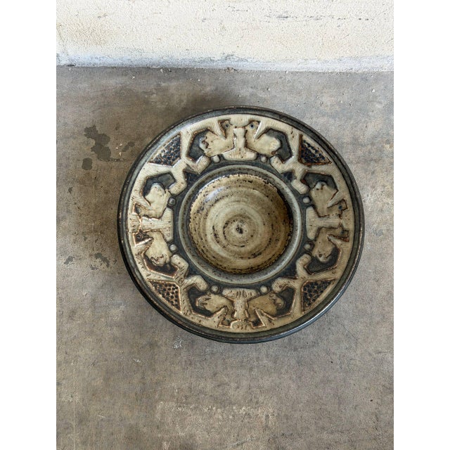 This vintage ceramic dish features a shallow circular basin with a wide decorative rim. The border showcases a series of...