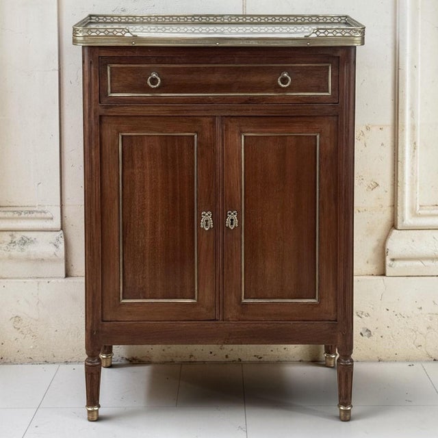 A French Louis XVI mahogany cabinet crowned with Carrara marble and a pierced brass gallery, glowing with the deep, warm...