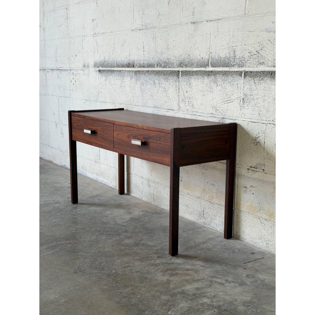 This minimalist hallway console table features a long rectilinear top flanked by slightly raised side lip borders. Two...