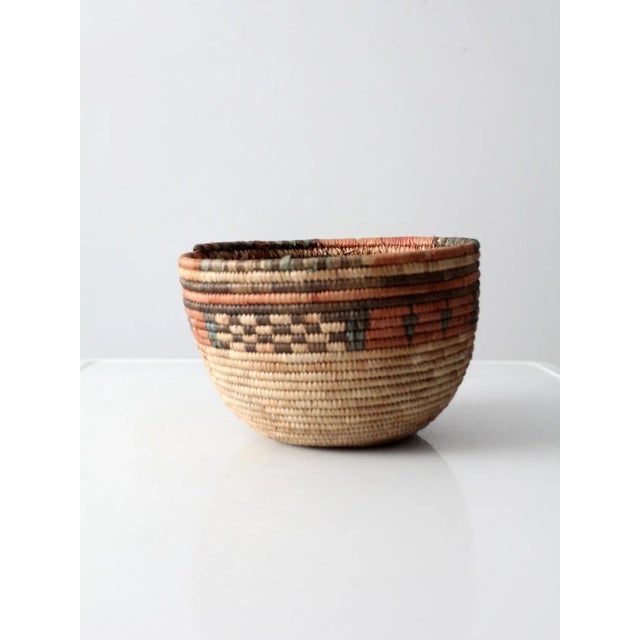 A beautifully woven vintage tribal basket featuring an intricate geometric pattern in soft red, green, and black tones....
