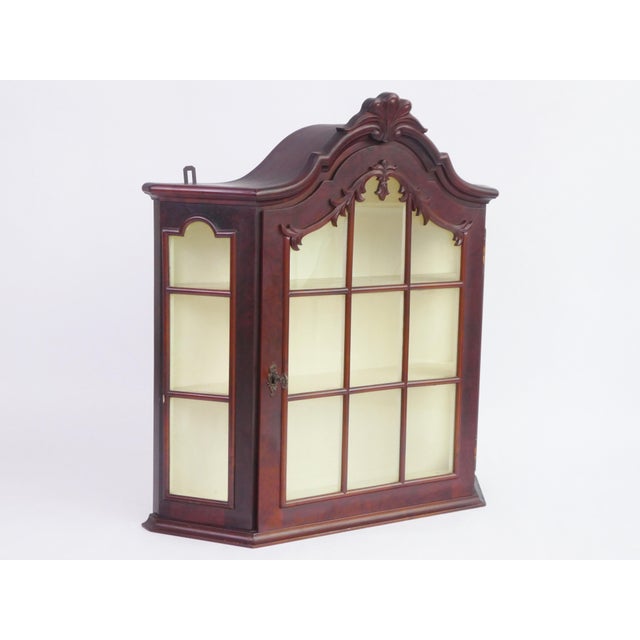 A mid century wooden wall display with glass door and white shelves in the inside. It has an amazing ornament on the top...