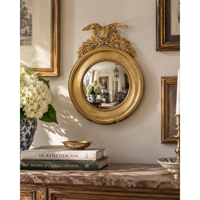 A Federal-style brass convex mirror crowned with a carved eagle, its wings outstretched above olive branches—an enduring...