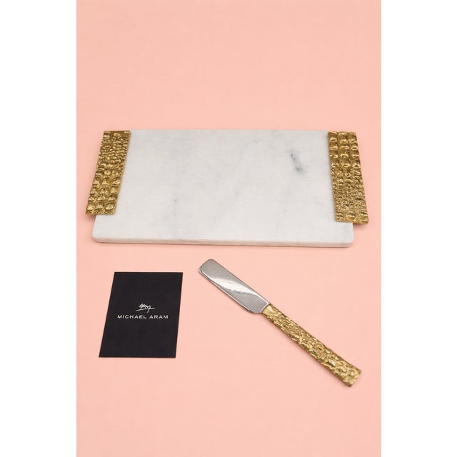 Michael Aram cheese board featuring a white marble surface with textured gold-tone handles from the Safari Collection. The...