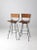 A pair of mid-century Arthur Umanoff style stools featuring black metal frames with woven rush backs and slatted wood...
