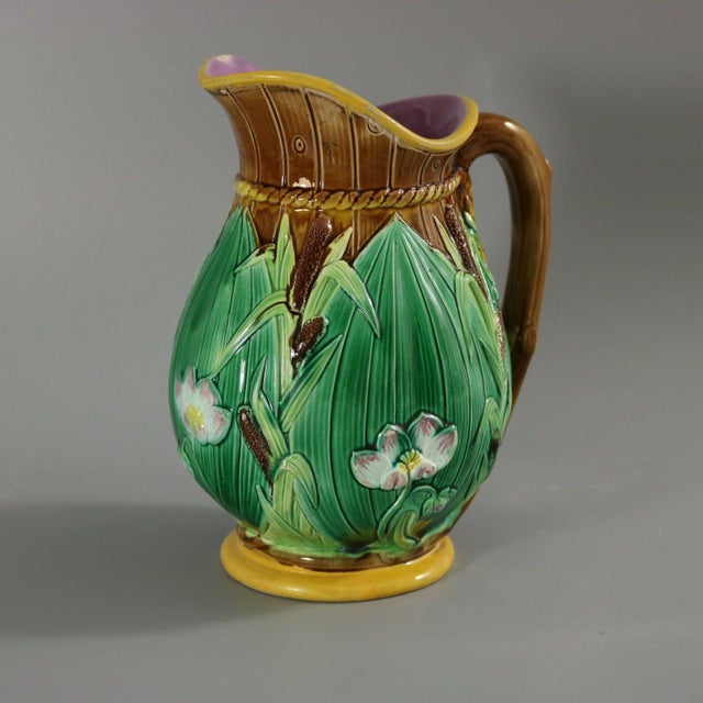 George Jones Majolica jug/pitcher which features lilies on a brown barrel ground. Colouration: brown, green, white, are...