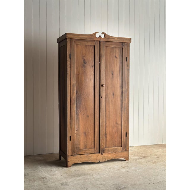 This antique pine cupboard cabinet features a tall, narrow silhouette with paneled doors, a hand-carved scroll crest, and...