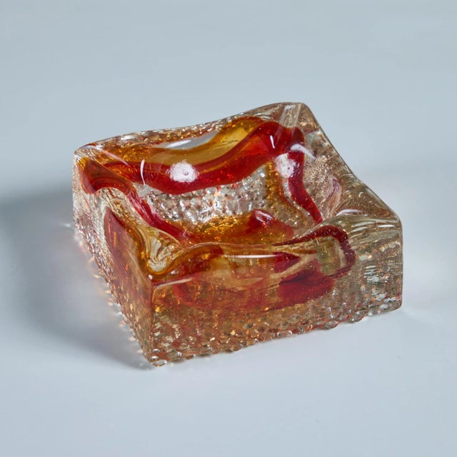 Mid-century ashtray features a square thick clear glass body with a reddish orange swirl throughout, a deep center well...