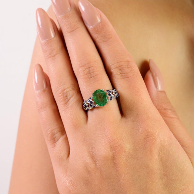 This beautiful 925 sterling silver ring features a vibrant 9x7 mm oval emerald paired with a sparkling 1.5 mm round blue...