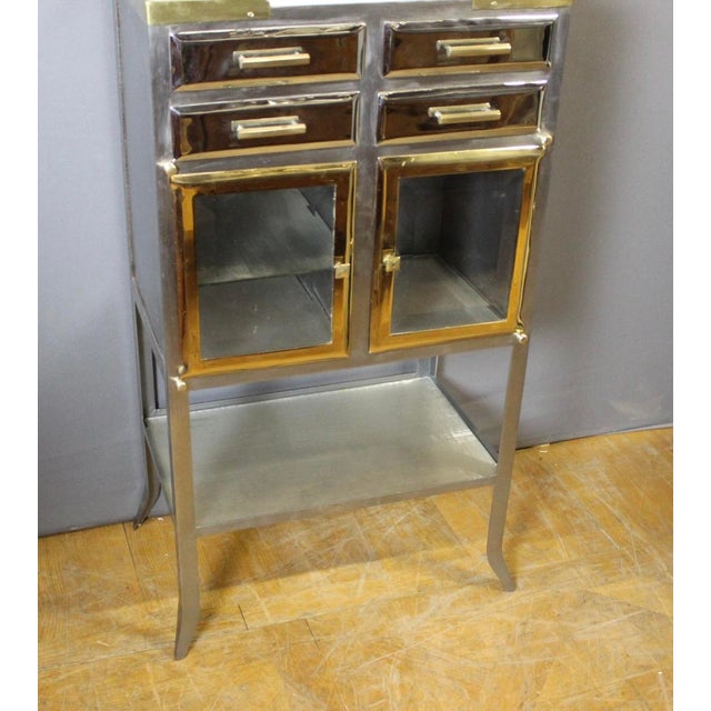 A dentist's cabinet in brushed, chromed, and gilded metal, featuring four drawers and two doors. The shelves are glass,...