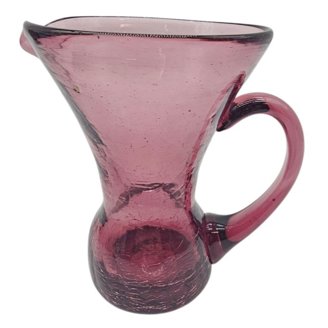 A soft plum ghost in glass, this pitcher carries the hush of handwork and time. Its neck curves inward as if to listen,...