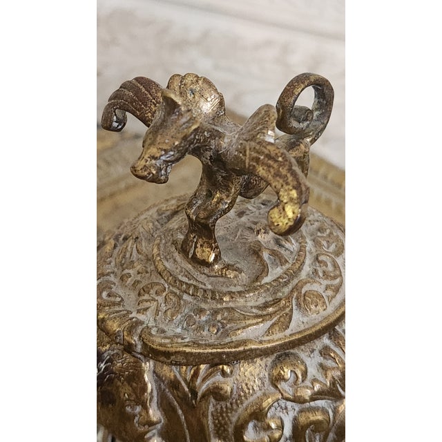 A decorative vintage brass inkwell tray featuring an ornate raised inkwell with a hinged lid, glass ink insert, and...