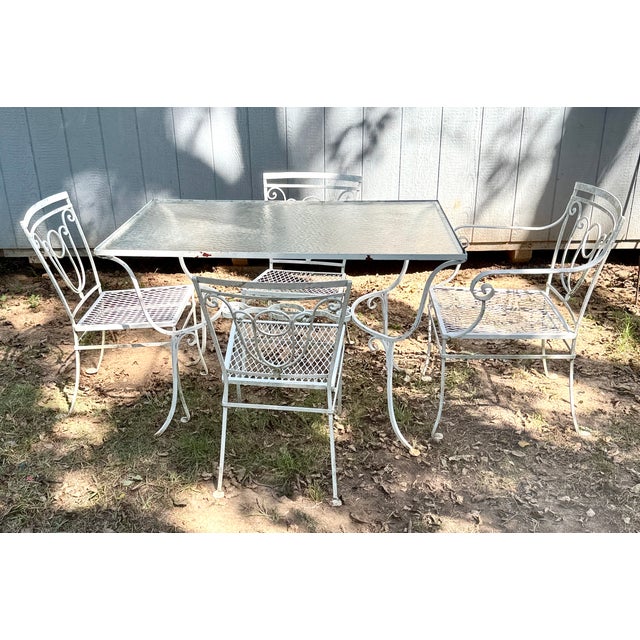This vintage white wrought iron patio dining set features a rectangular glass-top table paired with four matching mesh-...