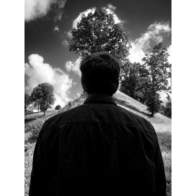 A solitary figure stands before a landscape alive with summer light. His face remains unseen, allowing him to become less...