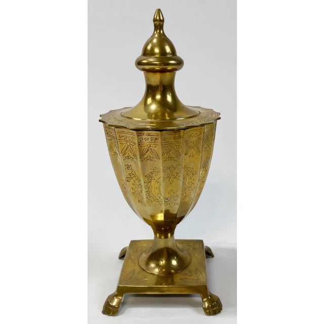 Vintage Neoclassical style scalloped lidded brass urn. Features intricate etchings and sits on a square footed base. Patina.