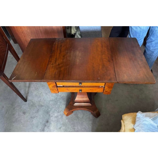 Empire-style sewing or work table featuring two drawers, hinged drop leaves, and a shaped pedestal supported by a platform...